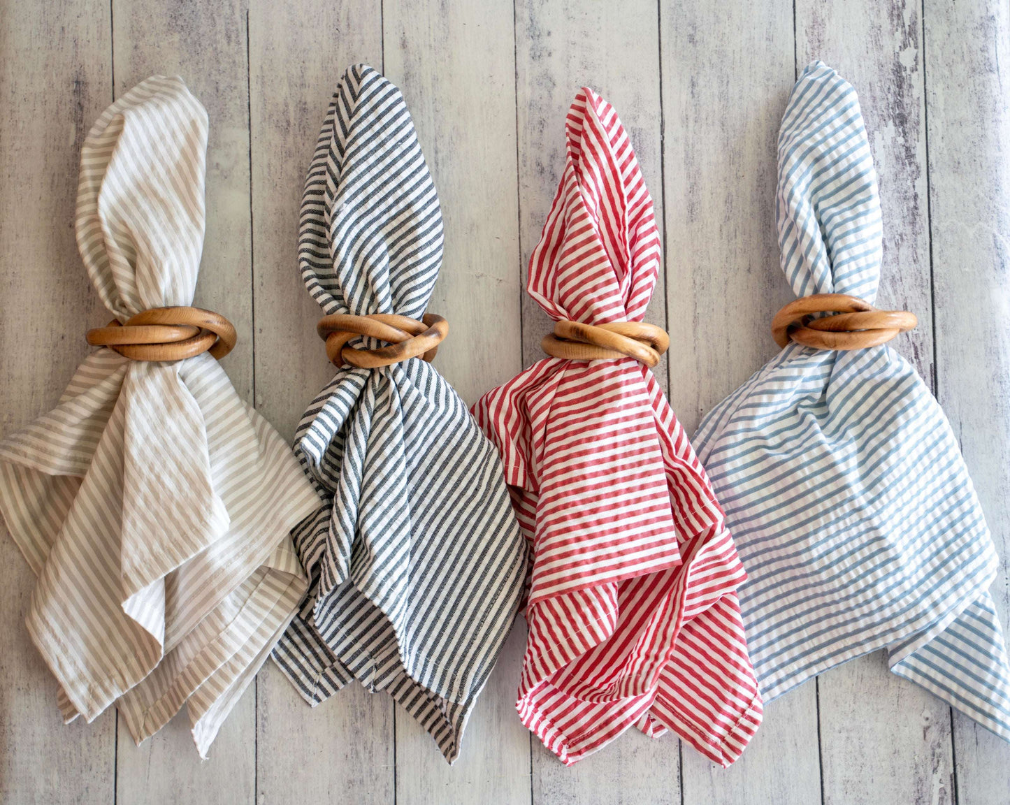 Four round wood napkin rings holding striped fabric napkins in tan, black, red, and blue on a light wood surface.