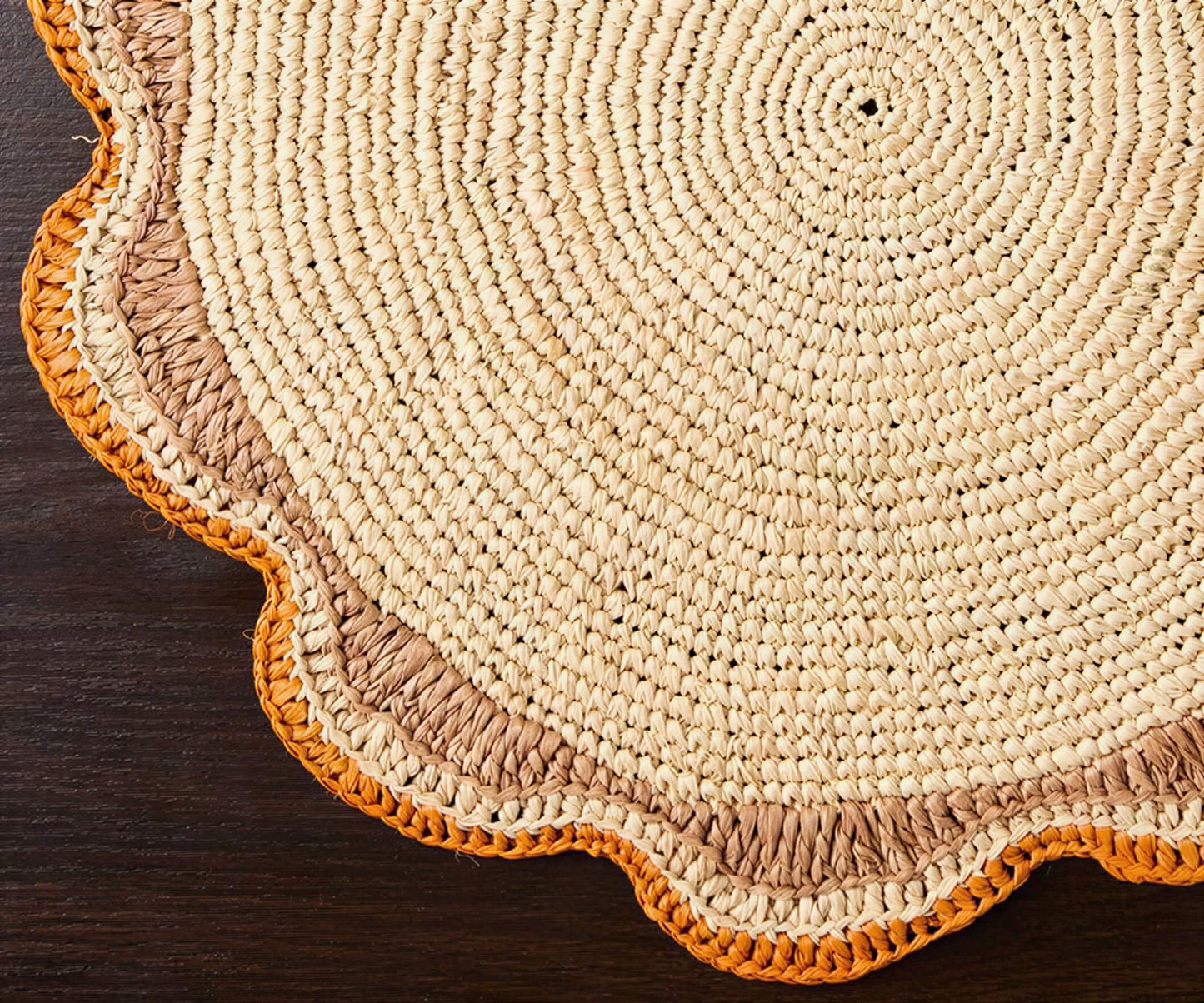Round raffia woven placemat with a scalloped edge, featuring natural fibers and an orange trim on a dark wooden surface.