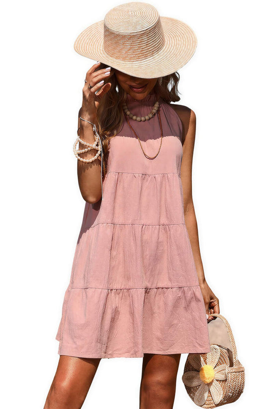 Halter neck cotton linen dress in light pink with tiered design, worn by a model holding a straw bag and wearing a wide-br...