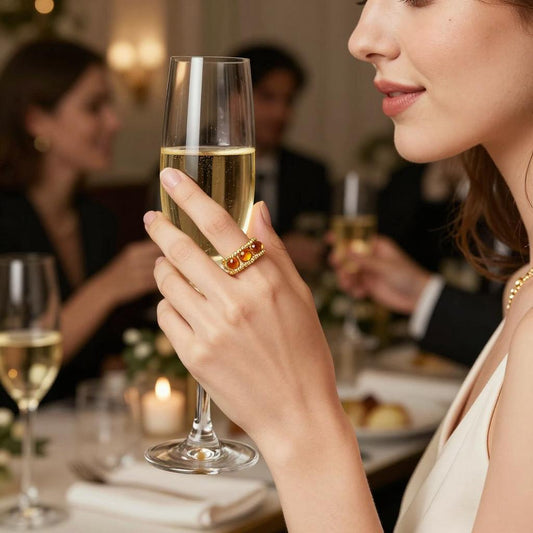 A young woman wearing an orange eternity ring holding a champagne glass with a group of people seated at a dinner table.