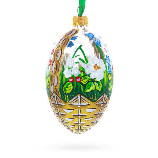 Egg-shaped glass ornament featuring a hand-painted gold basket filled with white and blue flowers and green leaves.