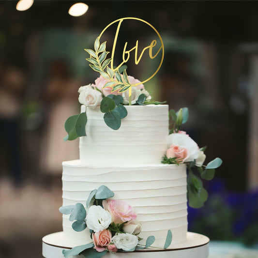 Gold acrylic "Love" wedding cake topper on a three-tiered white cake adorned with pink and white roses and greenery.