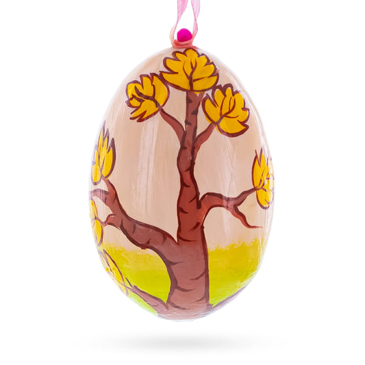 Oval wooden ornament painted with a brown tree and yellow leaves on a tan background, hanging from a pink ribbon.