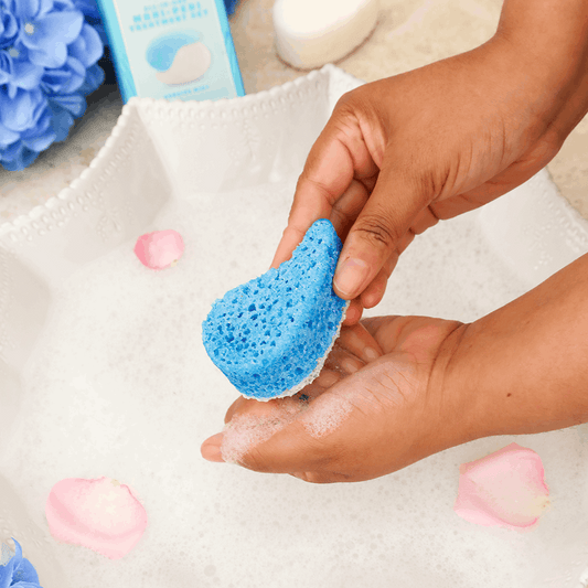 Hand holding a blue sponge with soap bubbles, surrounded by flowers and bath products.