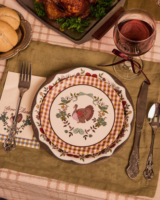 Stacked round paper plates with a turkey and floral design on a green placemat with silver cutlery and a wine glass.