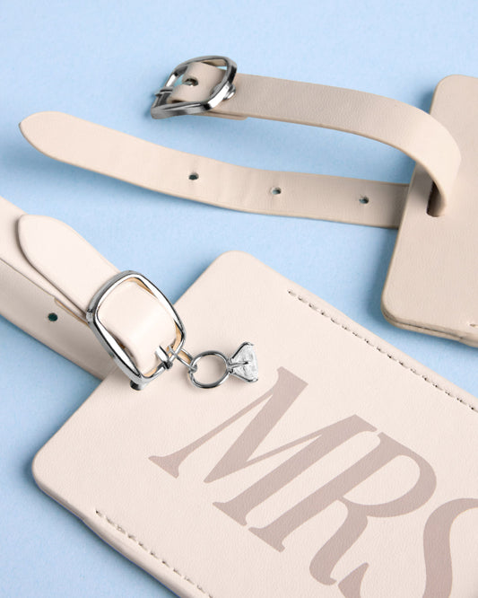 Two cream faux-leather luggage tags with silver buckles. One tag features a diamond ring charm and MRS printed in tan.