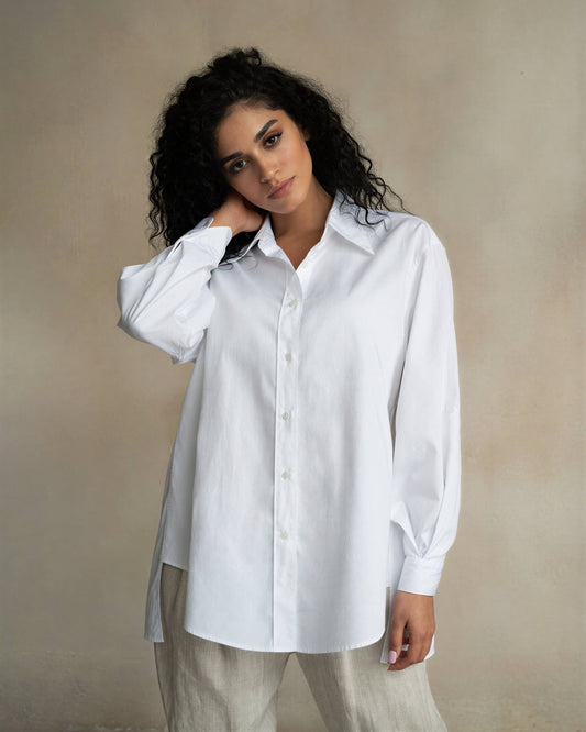 Woman wearing an oversized white cotton Terza shirt with a collar and long sleeves against a tan background.