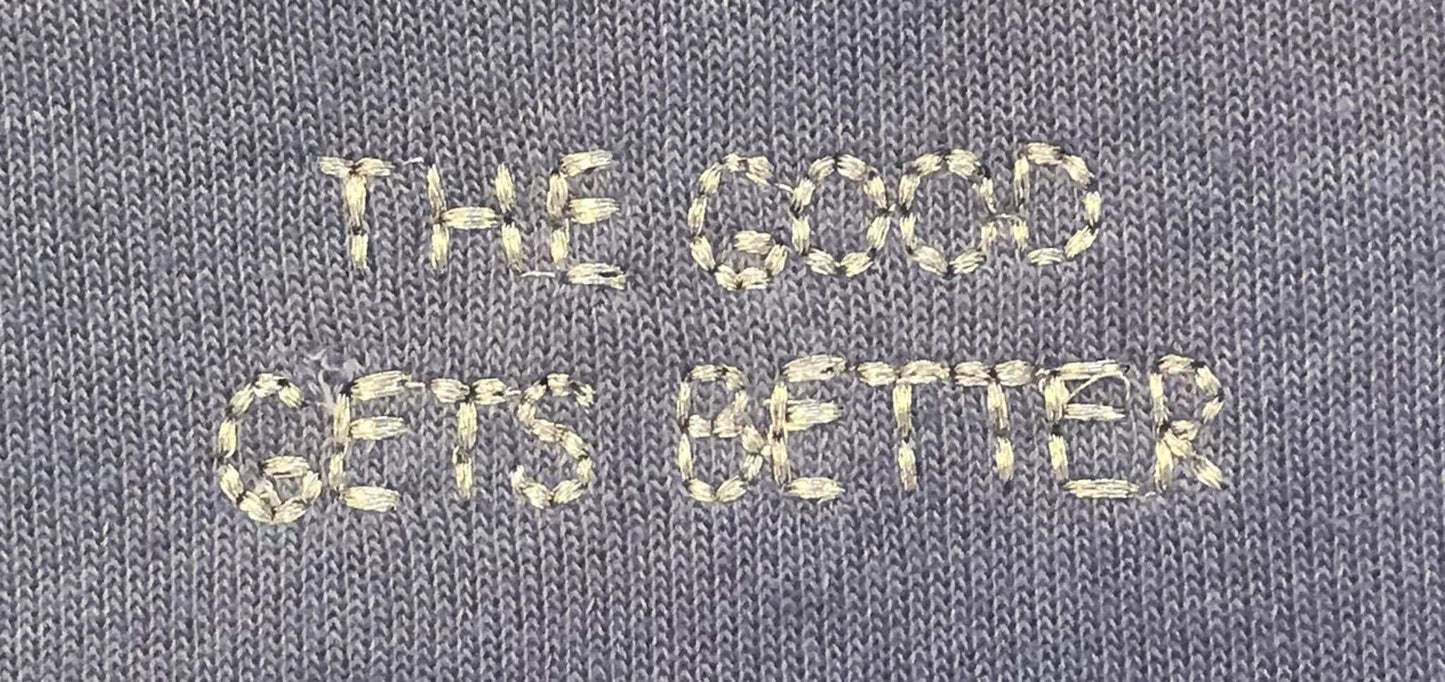 Close-up of a gray knit fabric with cream-colored embroidered text reading 'THE GOOD GETS BETTER'.