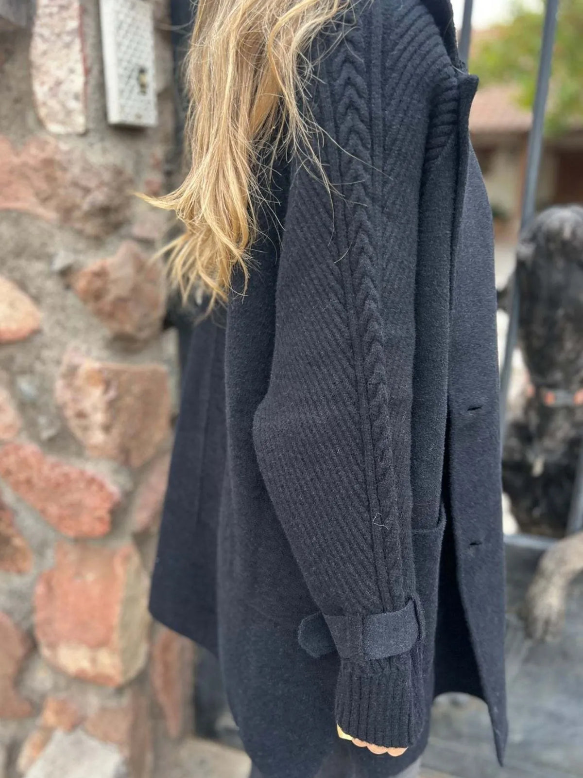 Model wearing a black double-breasted coat with cable-knit sleeves, buckled cuffs, wide lapels and front pockets.