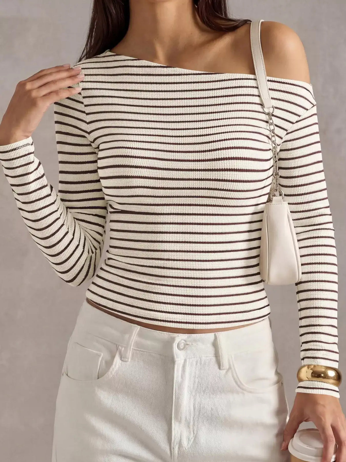 A woman wearing a fitted off-the-shoulder long-sleeve ribbed top with thin brown horizontal stripes on an off-white base, paired with white pants and a small off-white shoulder bag.