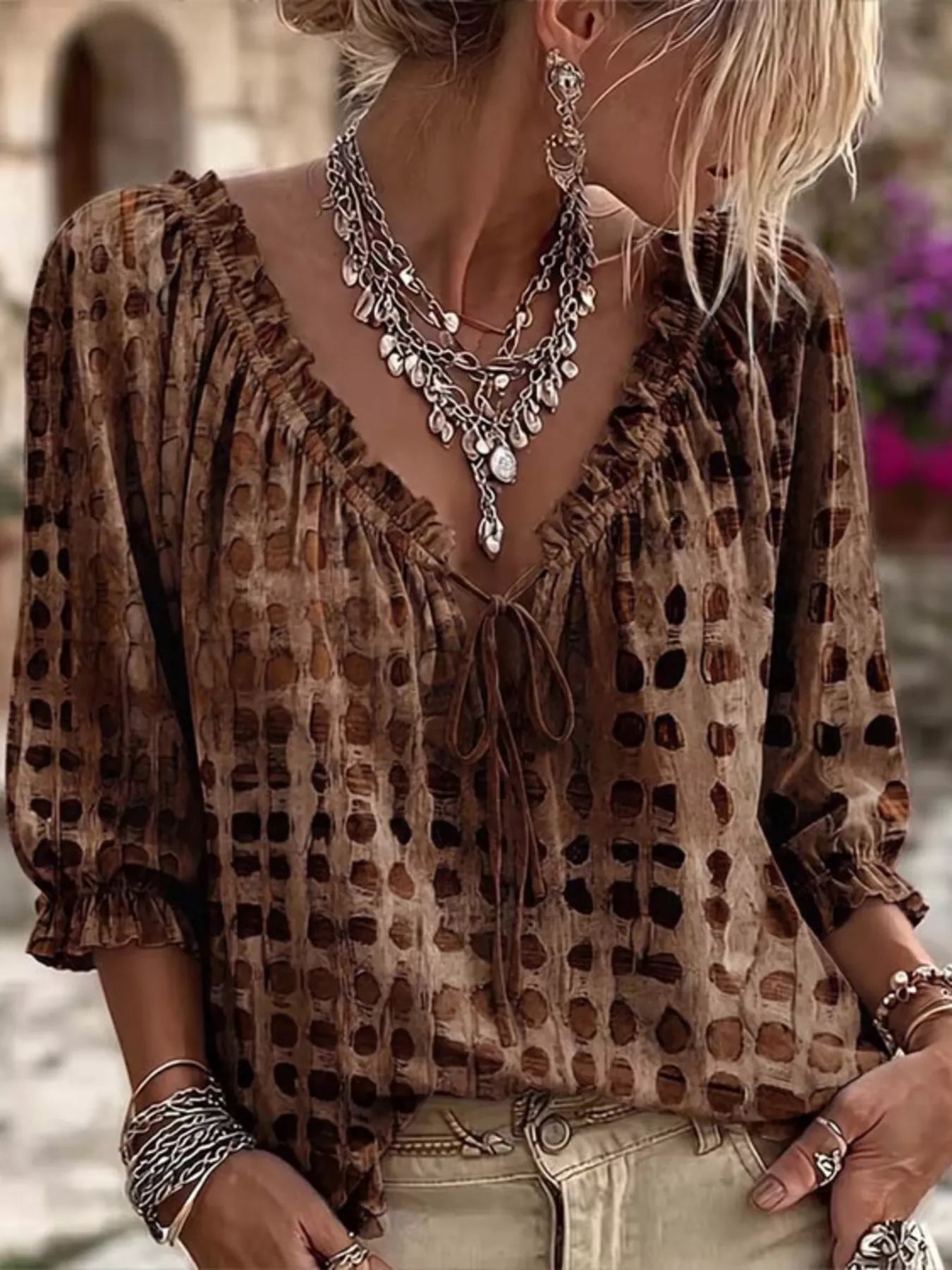 A woman wearing a brown and beige patterned blouse with a ruffled V-neckline and tie-front detail, styled with layered silver necklaces and bracelets.