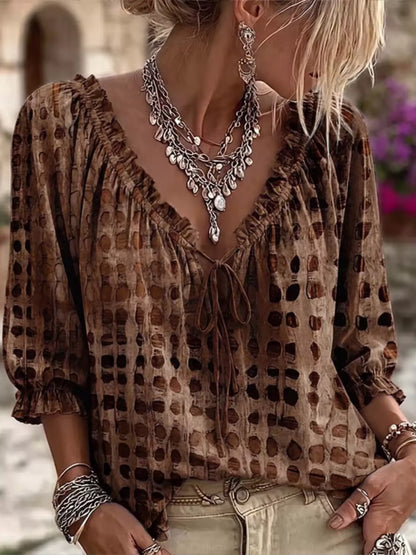 A woman wearing a brown and beige patterned blouse with a ruffled V-neckline and tie-front detail, styled with layered silver necklaces and bracelets.