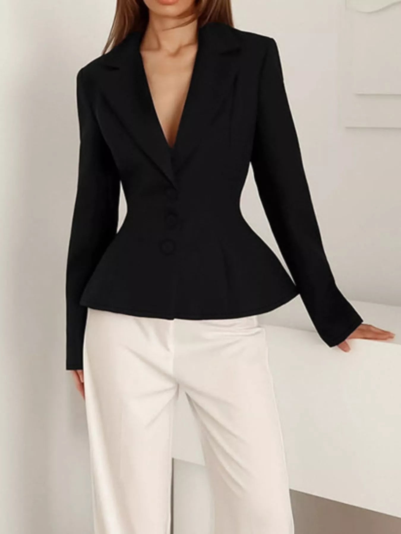 A woman wearing a fitted black long-sleeve blazer with a deep V-neck and peplum waist, paired with white wide-leg trousers, standing indoors against a light neutral wall.