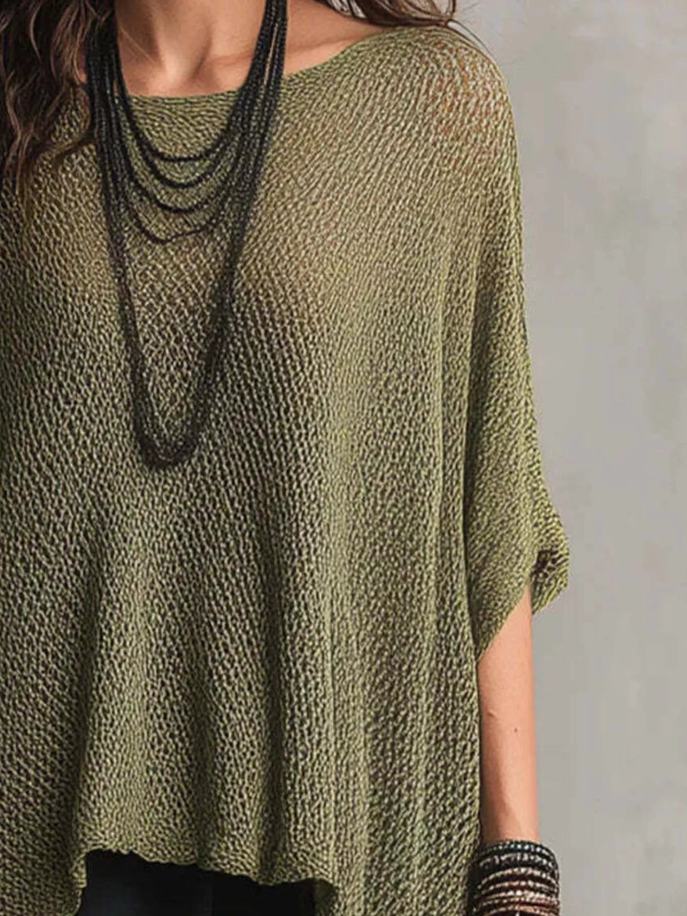 A woman wearing a loose, olive green knit top with a textured openwork pattern, styled with layered black necklaces and bracelets against a neutral gray background.