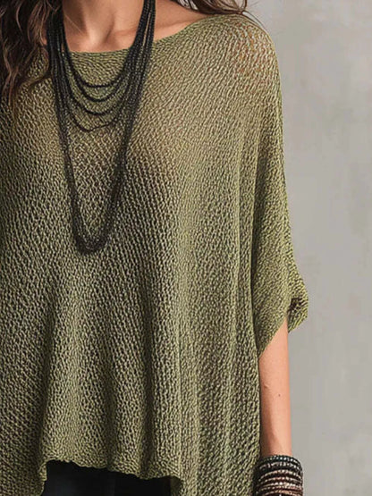 A woman wearing a loose, olive green knit top with a textured openwork pattern, styled with layered black necklaces and bracelets against a neutral gray background.