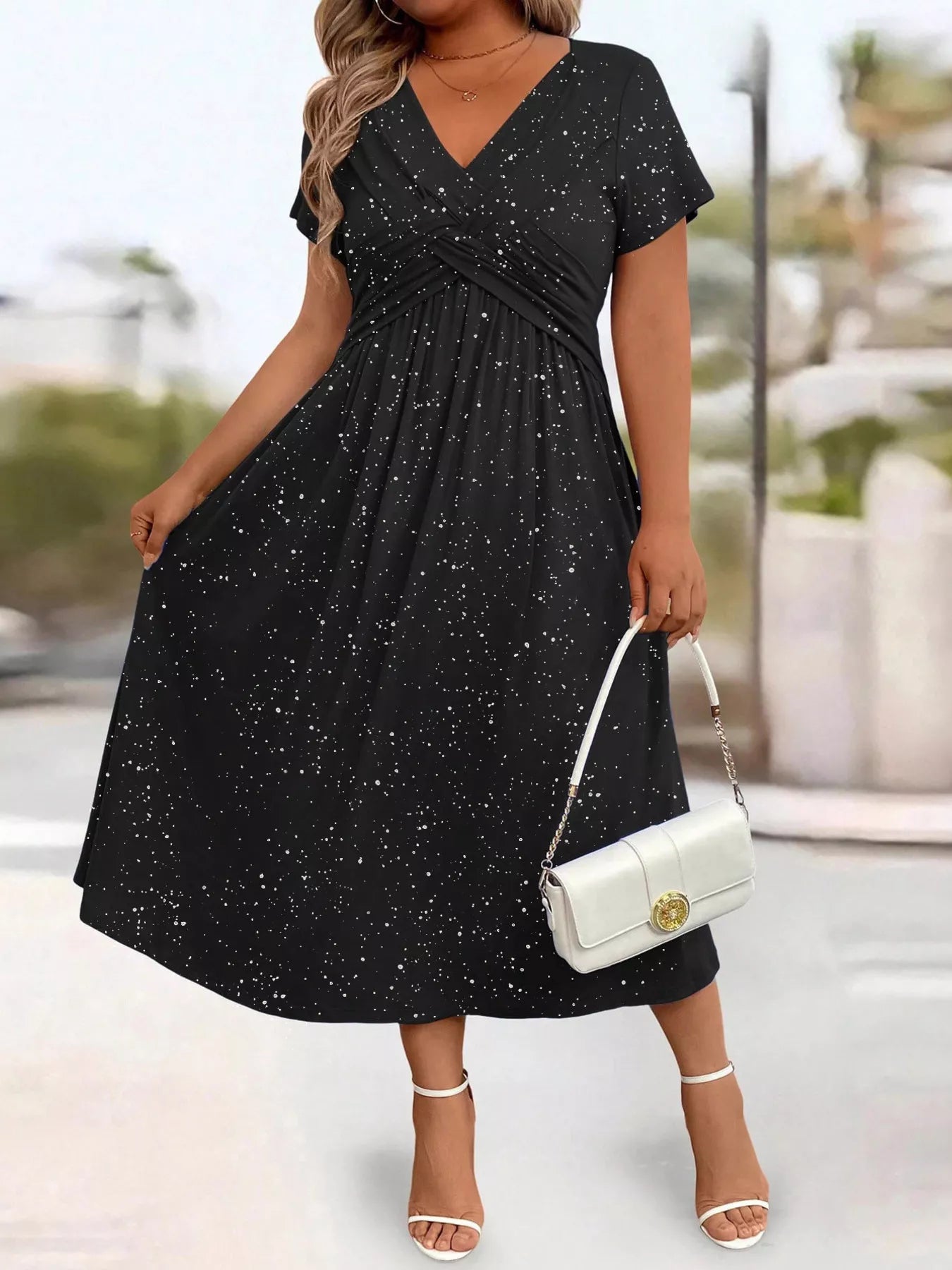 Woman wearing a black dress with white speckles, holding a white handbag, standing outdoors.