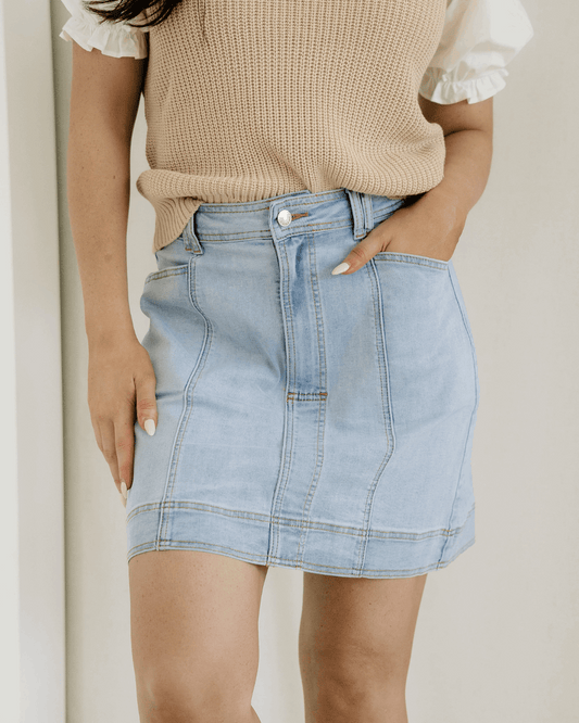 Person wearing a beige sweater and light blue denim skirt against a neutral background