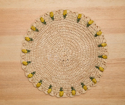 Round raffia crochet placemat featuring yellow tulip designs along the edge, set on a wooden surface.