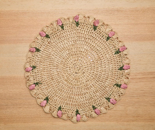 Hand-crocheted raffia palm placemat with pink tulip floral accents on a round design, set on a wooden surface.