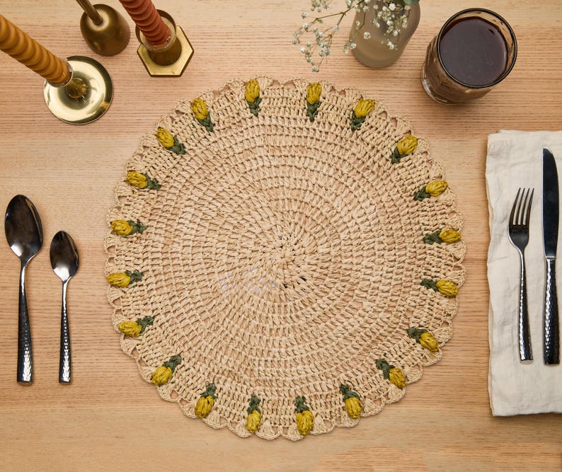 Handmade raffia crochet placemat with yellow tulip accents, set on a wooden table with dining utensils and decor.