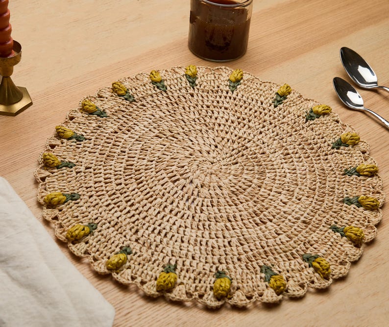Handmade raffia crochet placemat with yellow tulip accents, round shape, placed on a wooden table with utensils.