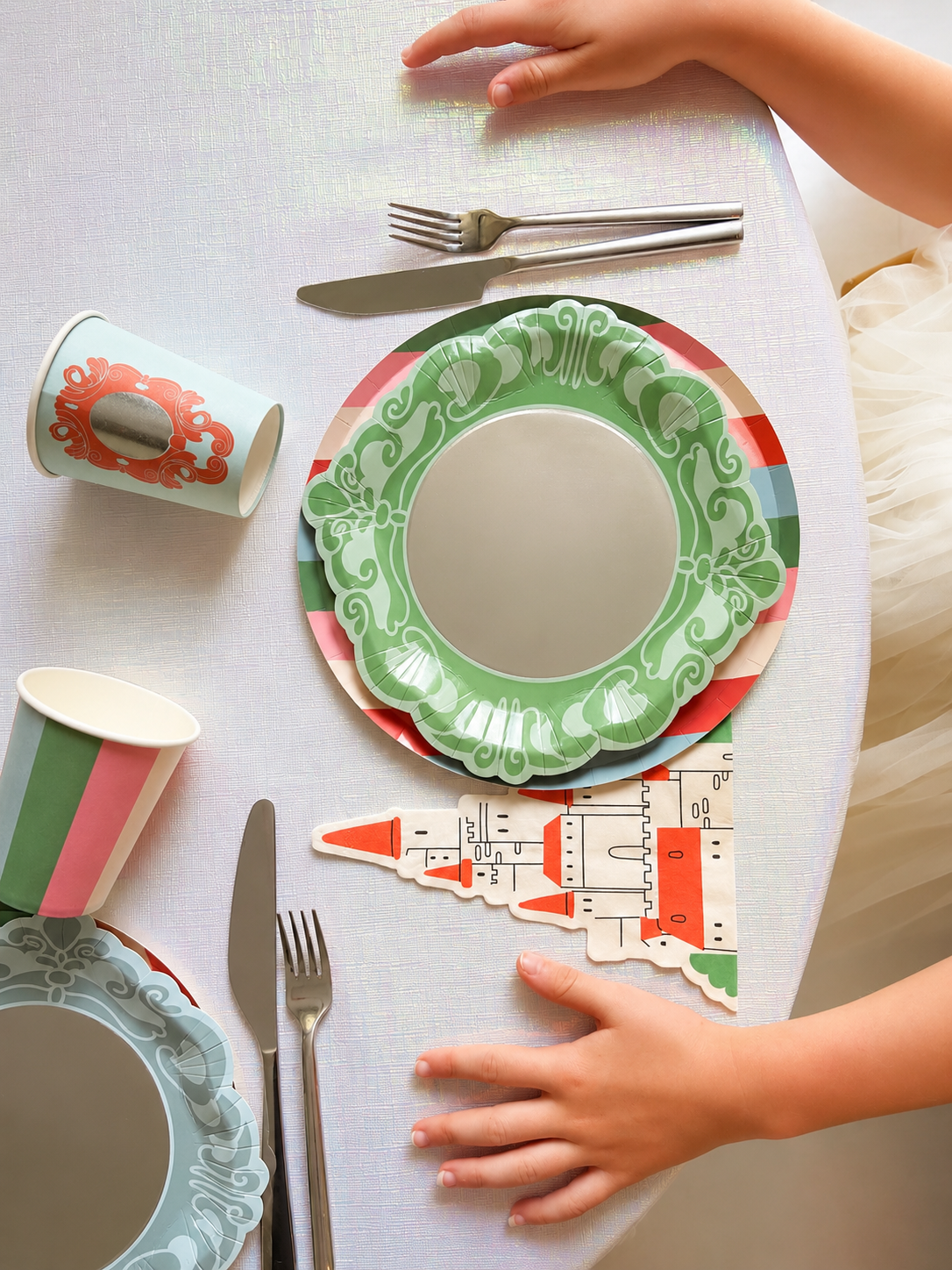 Green and blue scalloped paper plates, cups, and a castle-shaped napkin arranged on a white table with metal cutlery.