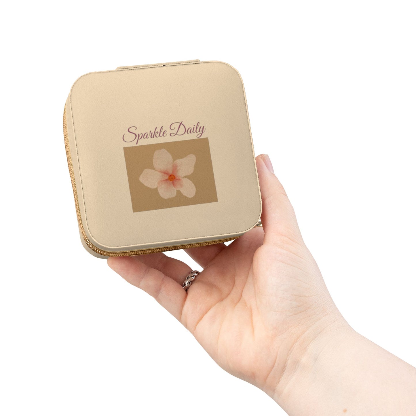 A hand holding a small square compact case with rounded corners. The case is a warm beige/champagne color with a printed brown square containing a white flower and the text 'Sparkle Daily'.