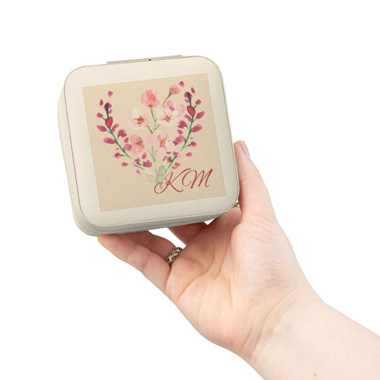 A hand holding a square cream-colored compact case with rounded corners. The front features a beige panel decorated with a pink and red floral heart design and the initials 'KM' in red script.