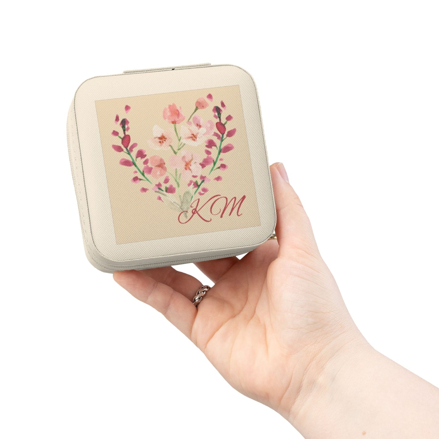 A hand holding a square cream-colored compact case with rounded corners. The front features a beige panel decorated with a pink and red floral heart design and the initials 'KM' in red script.