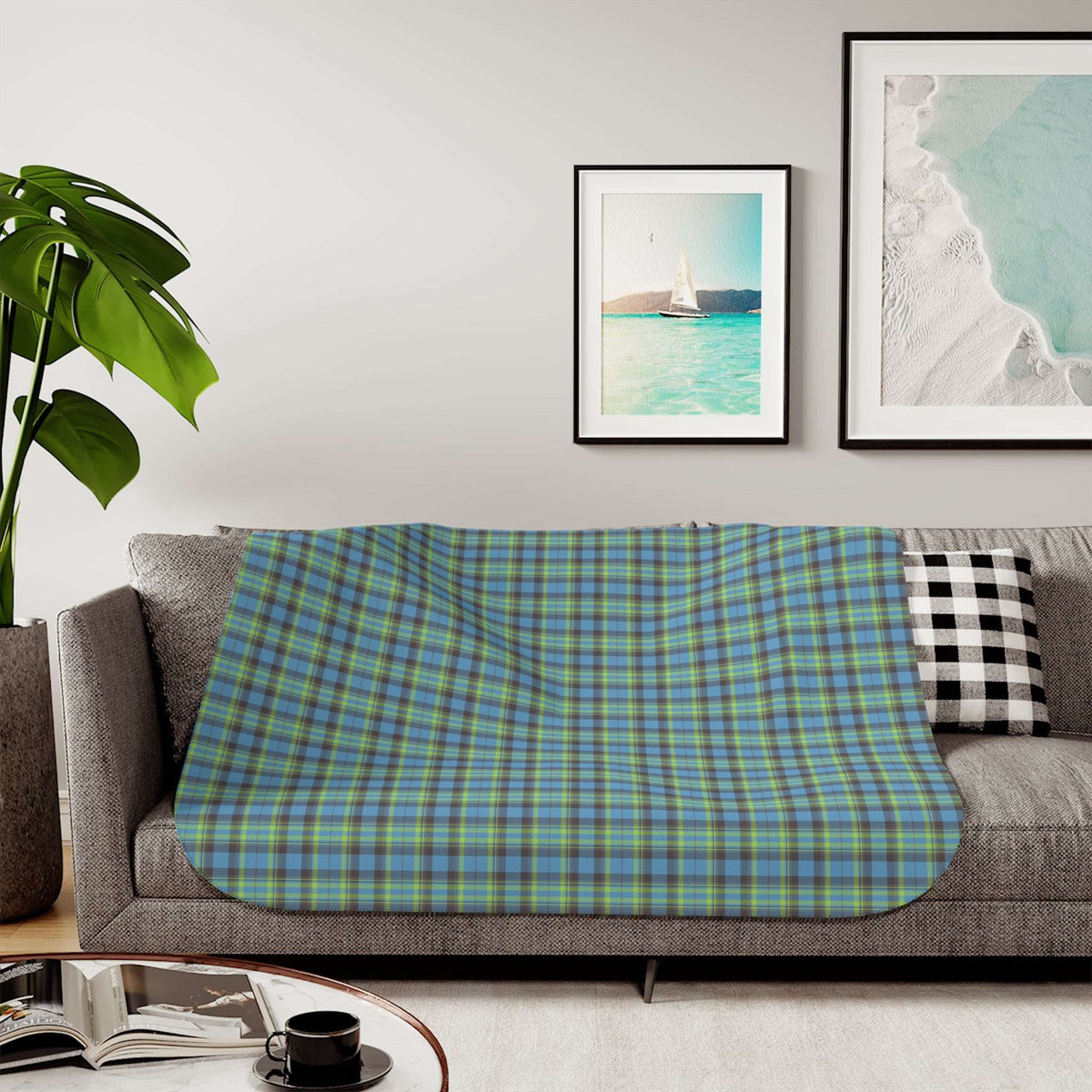 A plaid throw blanket draped over a gray sofa in a living room setting, featuring a blue, green, and yellow tartan pattern. The background includes framed wall art, a potted plant, and a coffee table with an open book and a black cup.