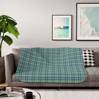 A plaid throw blanket draped over a gray sofa in a living room setting, featuring a blue, green, and yellow tartan pattern. The background includes framed wall art, a potted plant, and a coffee table with an open book and a black cup.