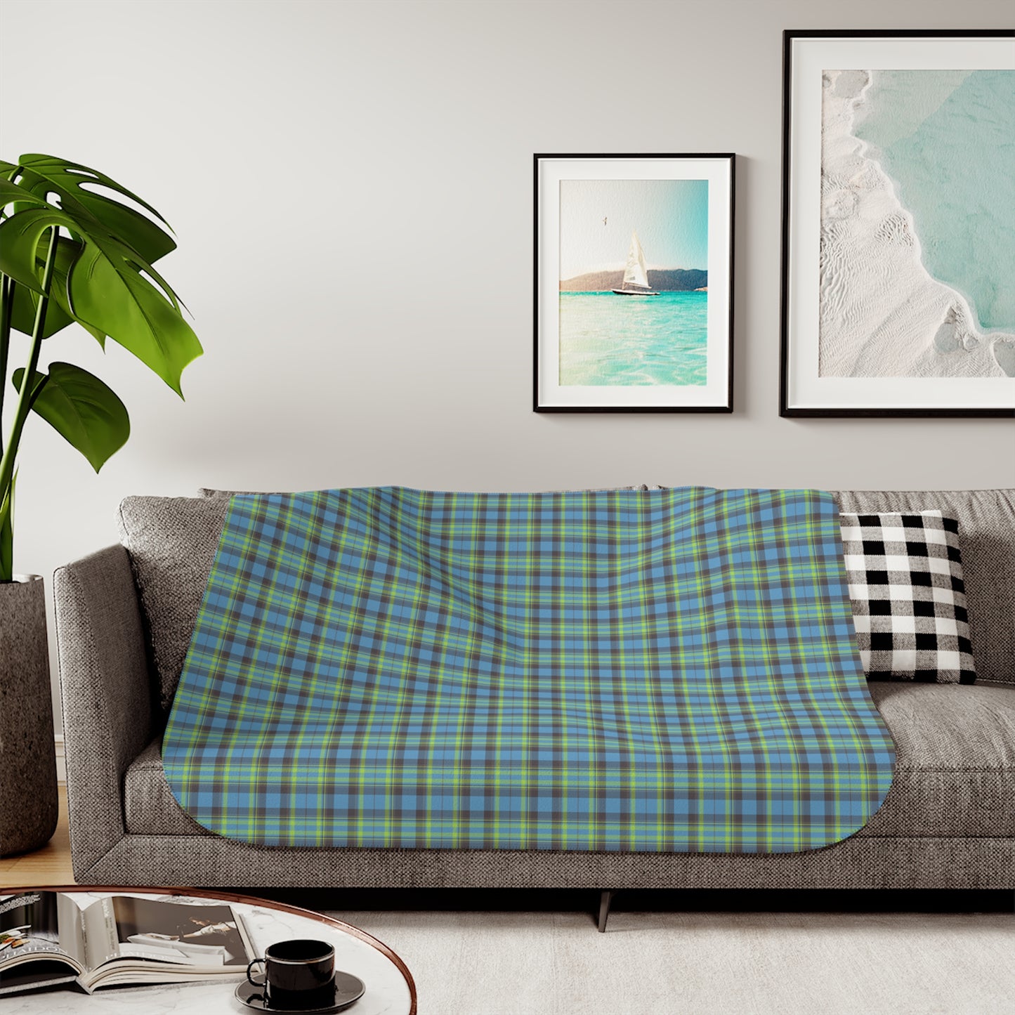 A plaid throw blanket draped over a gray sofa in a living room setting, featuring a blue, green, and yellow tartan pattern. The background includes framed wall art, a potted plant, and a coffee table with an open book and a black cup.
