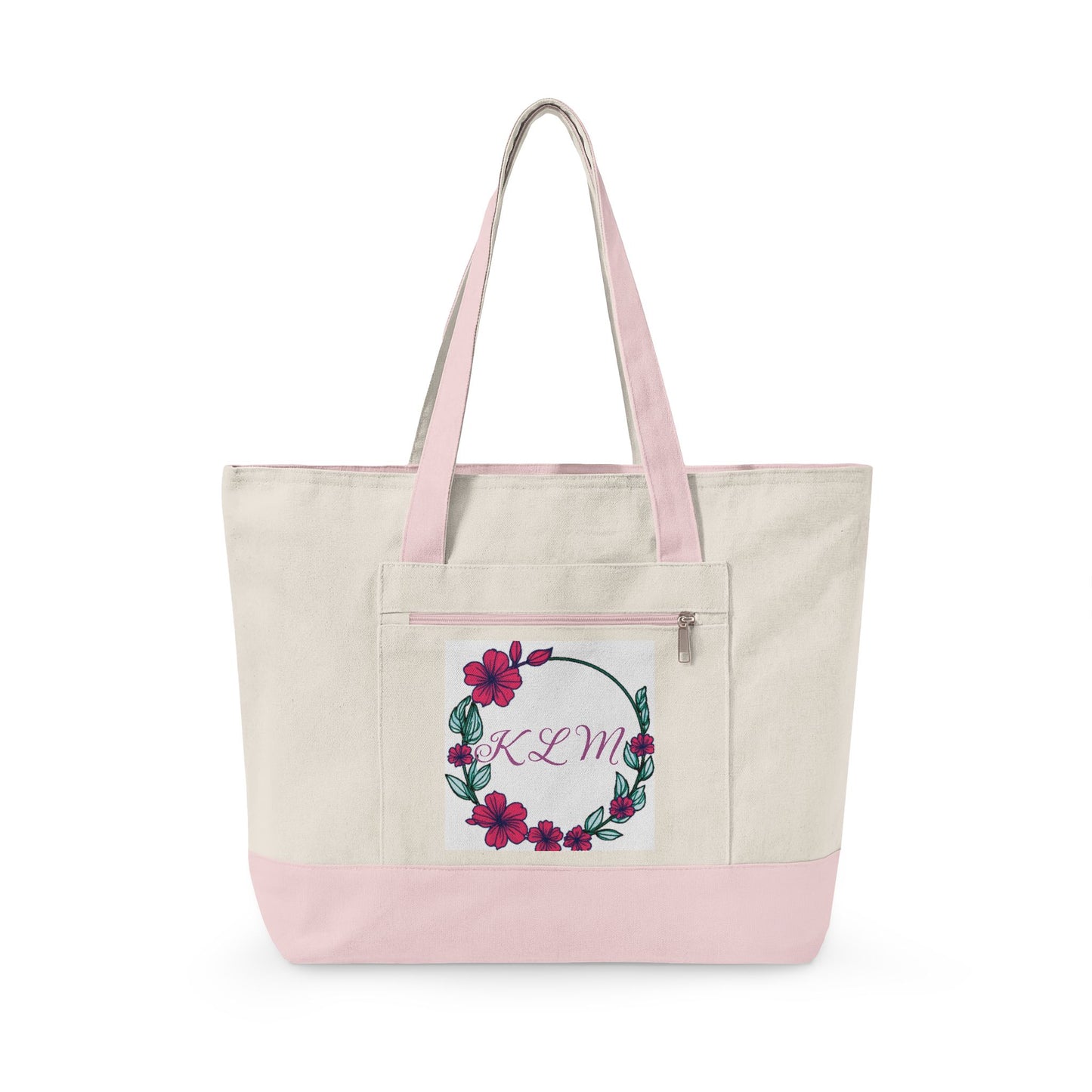 A canvas tote bag with a natural beige body, light pink handles and bottom base, featuring a front pocket with a floral wreath design and the monogram 'KDM' in pink script.