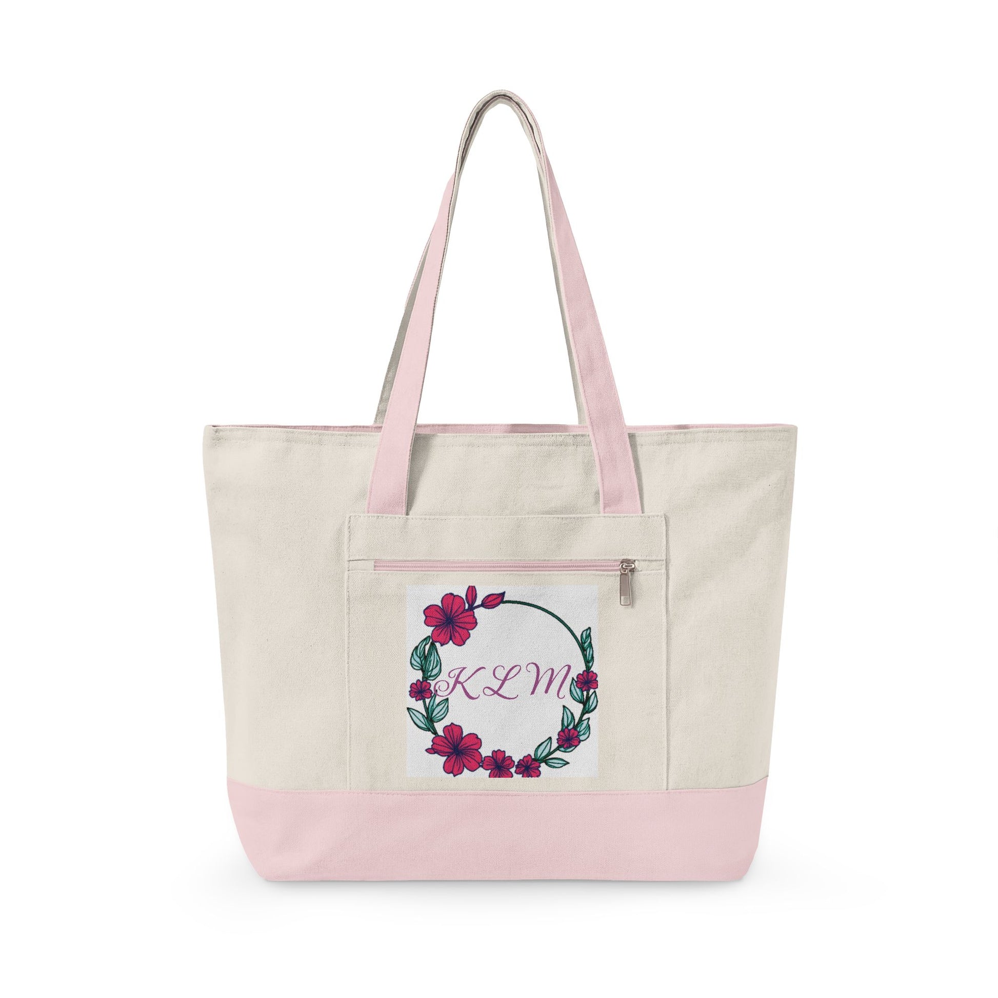 A canvas tote bag with a natural beige body, light pink handles and bottom base, featuring a front pocket with a floral wreath design and the monogram 'KDM' in pink script.