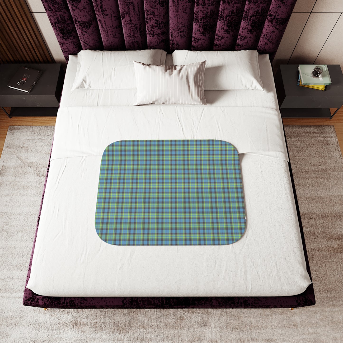 A top-down view of a bed with white bedding and a purple tufted headboard, featuring a rectangular blue and green plaid blanket with rounded corners laid across the lower half.