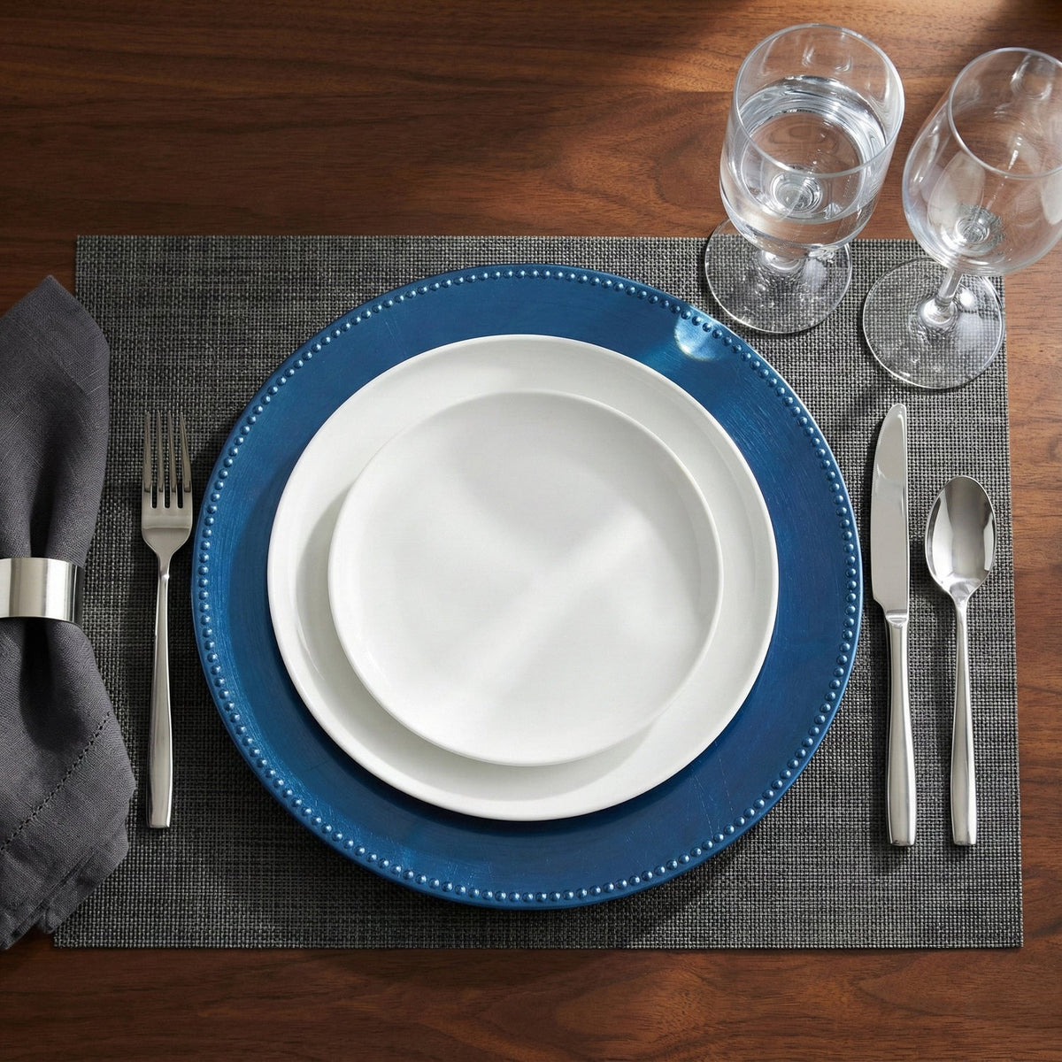 Dining table setting with blue plates, white bowls, silverware, and glasses on a wooden table.