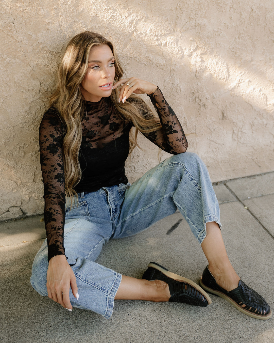 Woman sitting on a sidewalk wearing a black sheer top and blue jeans.