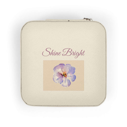 A square cream-colored zippered pouch with rounded corners, featuring the text 'Shine Bright' in a dusty rose script above a central square graphic of a purple and blue flower on a beige background.