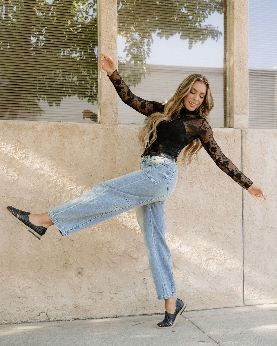 Woman in black lace top and blue jeans posing against a stone wall.