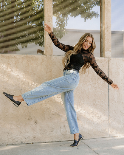 Woman in black lace top and blue jeans posing against a stone wall.