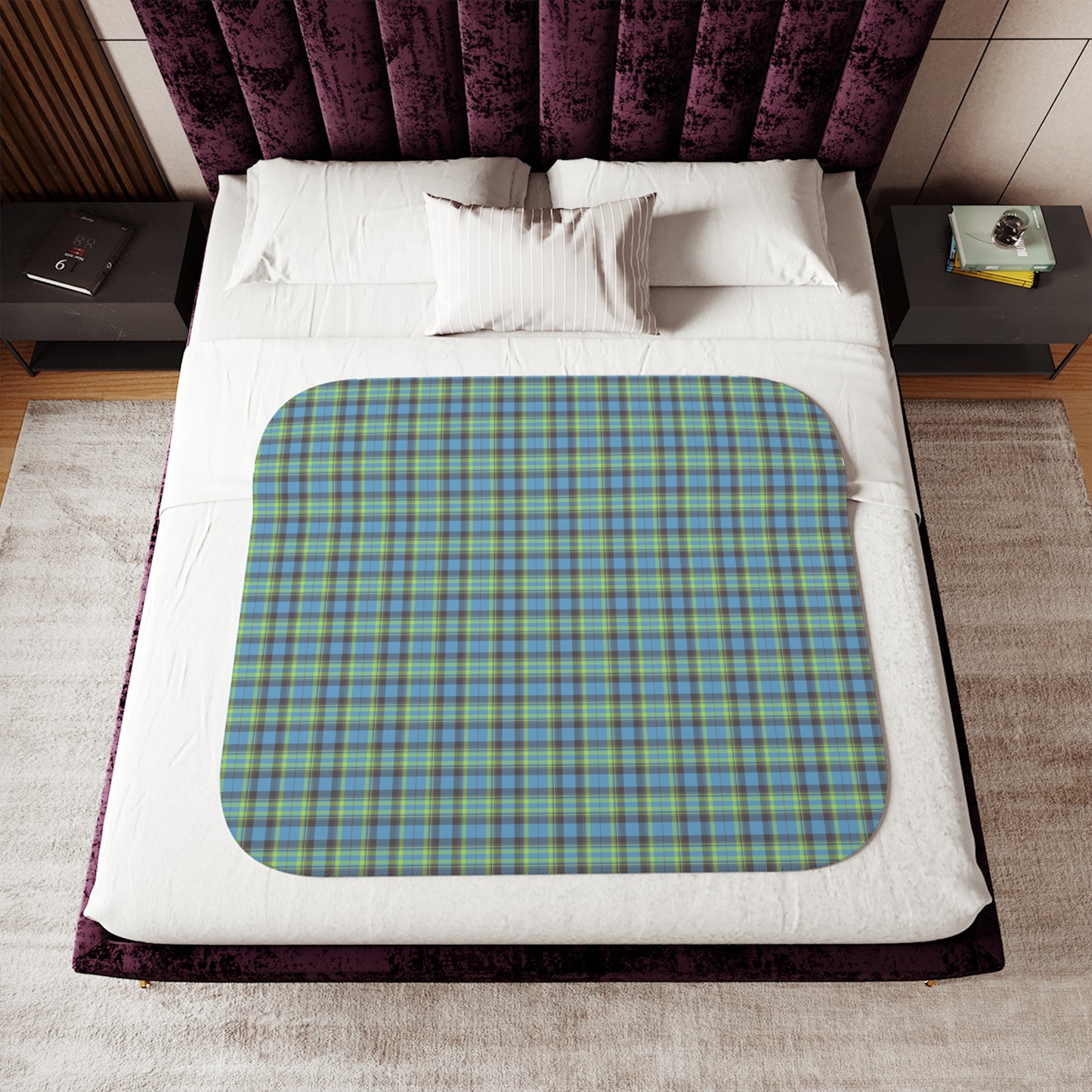 A square bath mat with rounded corners featuring a green, blue, and purple tartan plaid pattern, styled on a white bed with a purple velvet headboard.