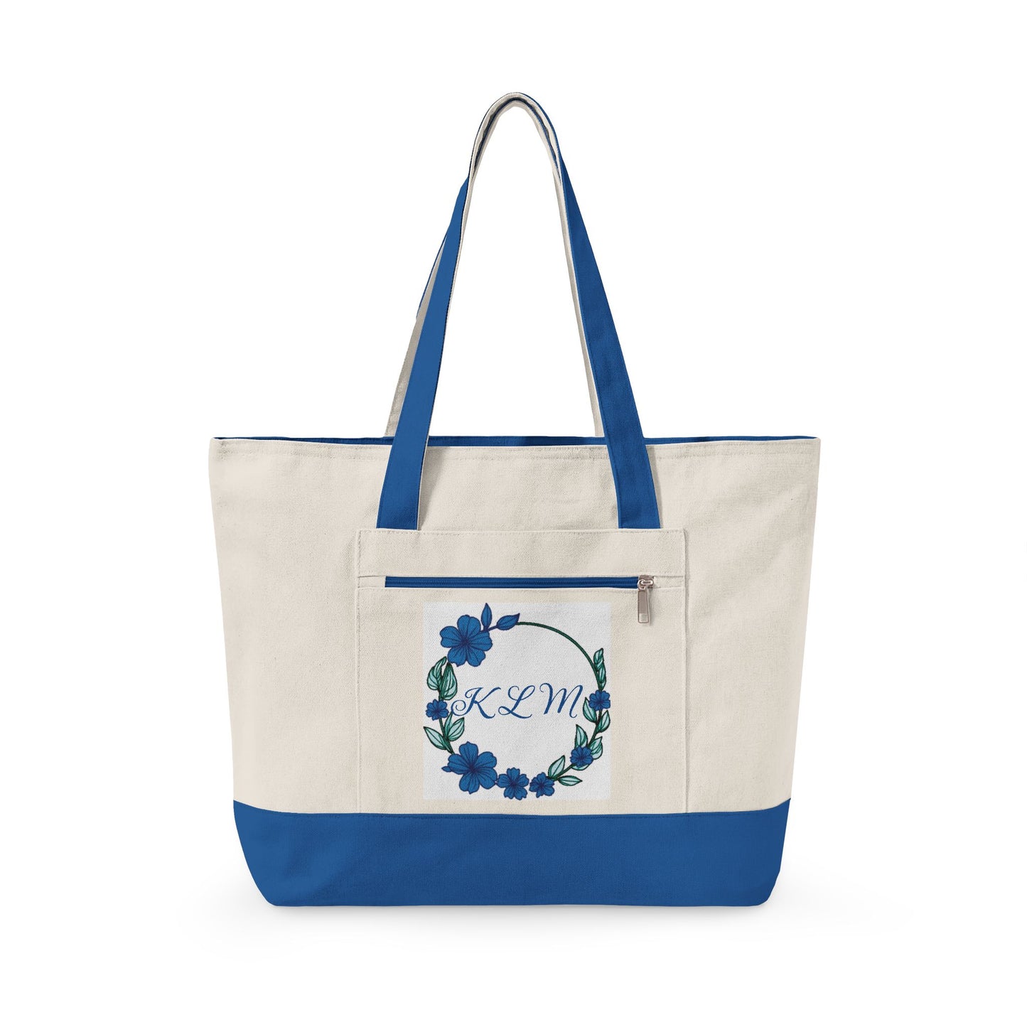 An off-white canvas tote bag with royal blue handles and bottom trim, featuring a front pocket with a blue floral wreath design and the monogram 'KCM'.