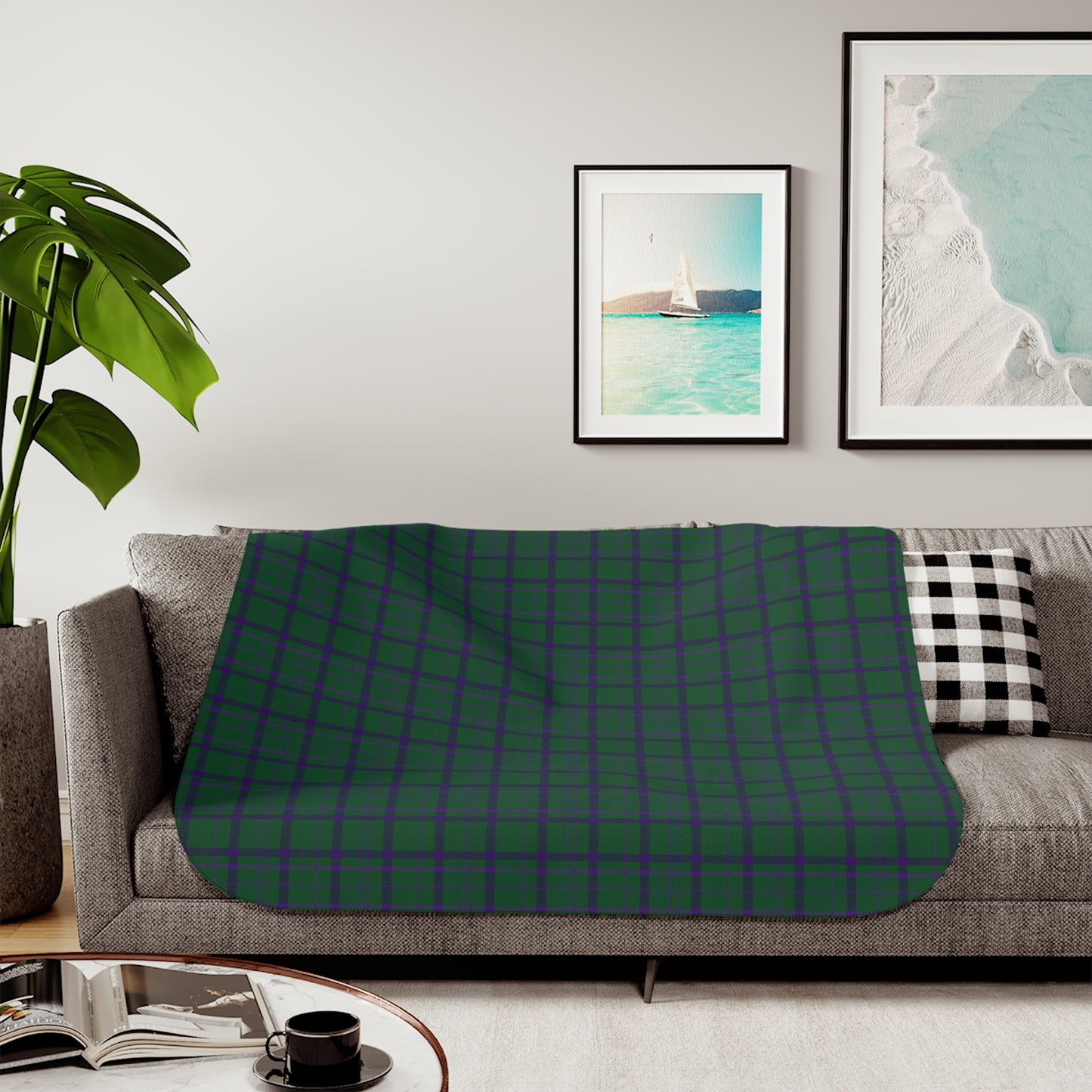 A green and purple tartan plaid throw blanket is draped over a gray fabric sofa in a modern living room setting. The room features a light gray wall with two framed art prints, a potted green plant on the left, and a black and white checkered pillow on the right.