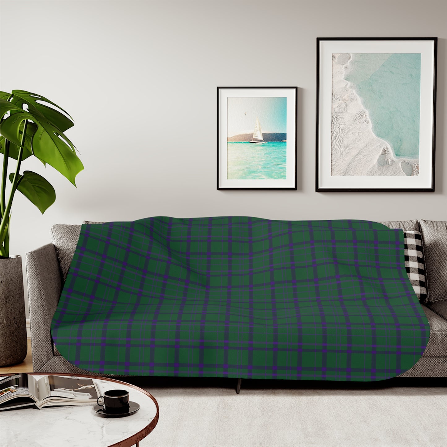 A green and purple tartan plaid throw blanket is draped over a gray sofa in a modern living room setting. The room features a light gray wall with two framed art prints, a potted green plant on the left, and a round coffee table in the foreground.