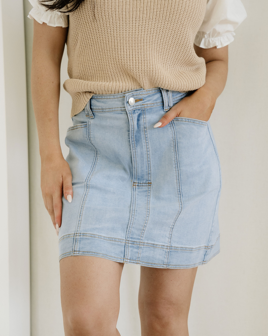 Person wearing a beige sweater and light blue denim skirt against a neutral background