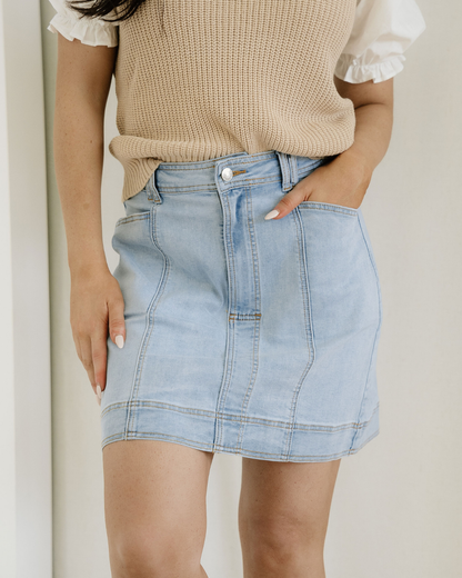 Person wearing a beige sweater and light blue denim skirt against a neutral background