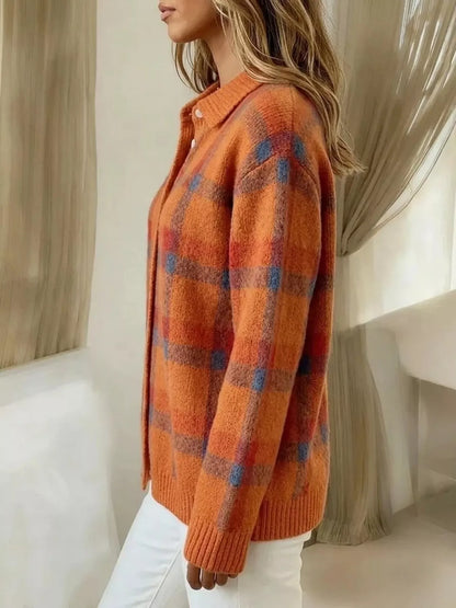 A woman wearing a fuzzy orange knit cardigan with a blue and red plaid pattern, paired with white pants, shown in a softly lit indoor setting.