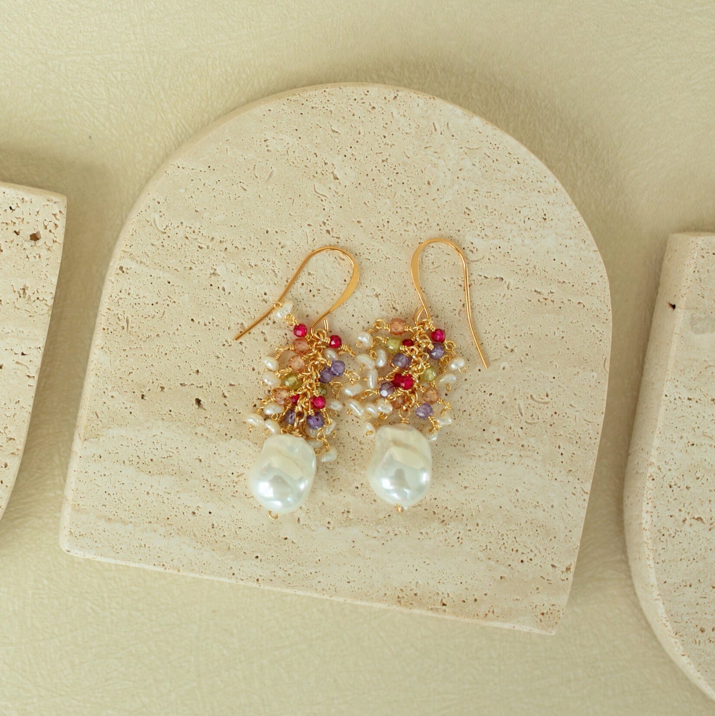 Gold earrings with pearls and red stones on a beige stone surface