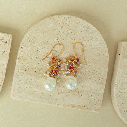 Gold earrings with pearls and red stones on a beige stone surface
