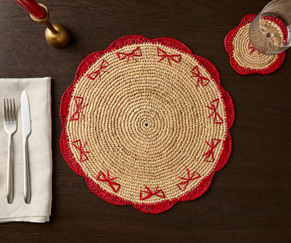Handwoven raffia fringe coaster with red scalloped edges and bow designs on a dark wooden table setting.